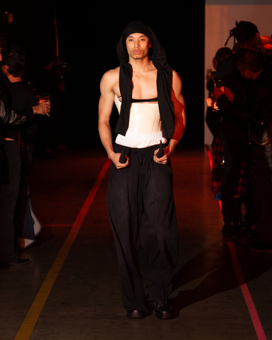 Slender male model with tan skin wearing a black head wrap, white corset and black pants walking on a runway against a dark backdrop